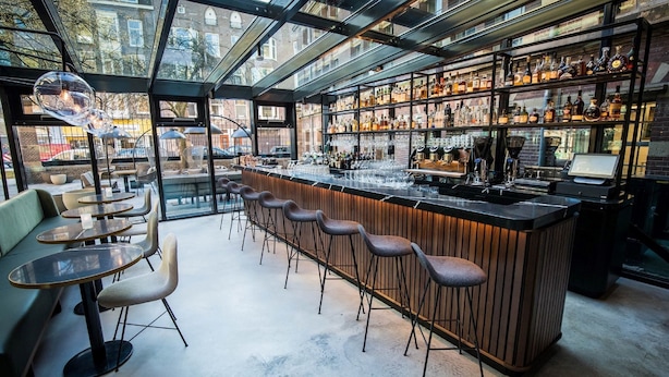 A modern bar with a glass ceiling, bar stools, and a variety of liquor bottles. - Pestana Amsterdam Riverside (Amsterdam)