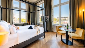 A hotel room with a large bed, a desk with chairs, a view of the city, and a minibar. - Pestana Amsterdam Riverside (Amsterdam)