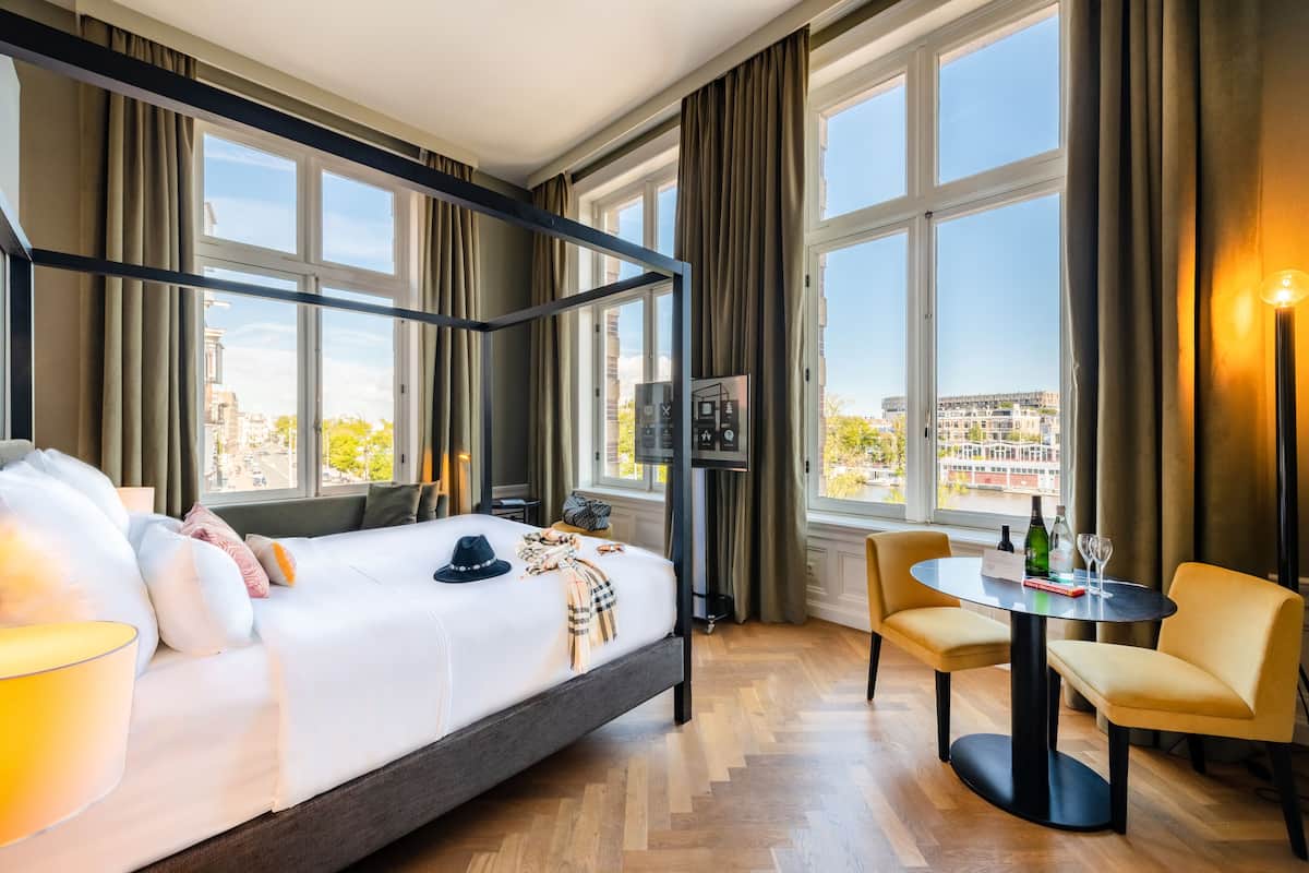 A hotel room with a large bed, a desk with chairs, a view of the city, and a minibar.