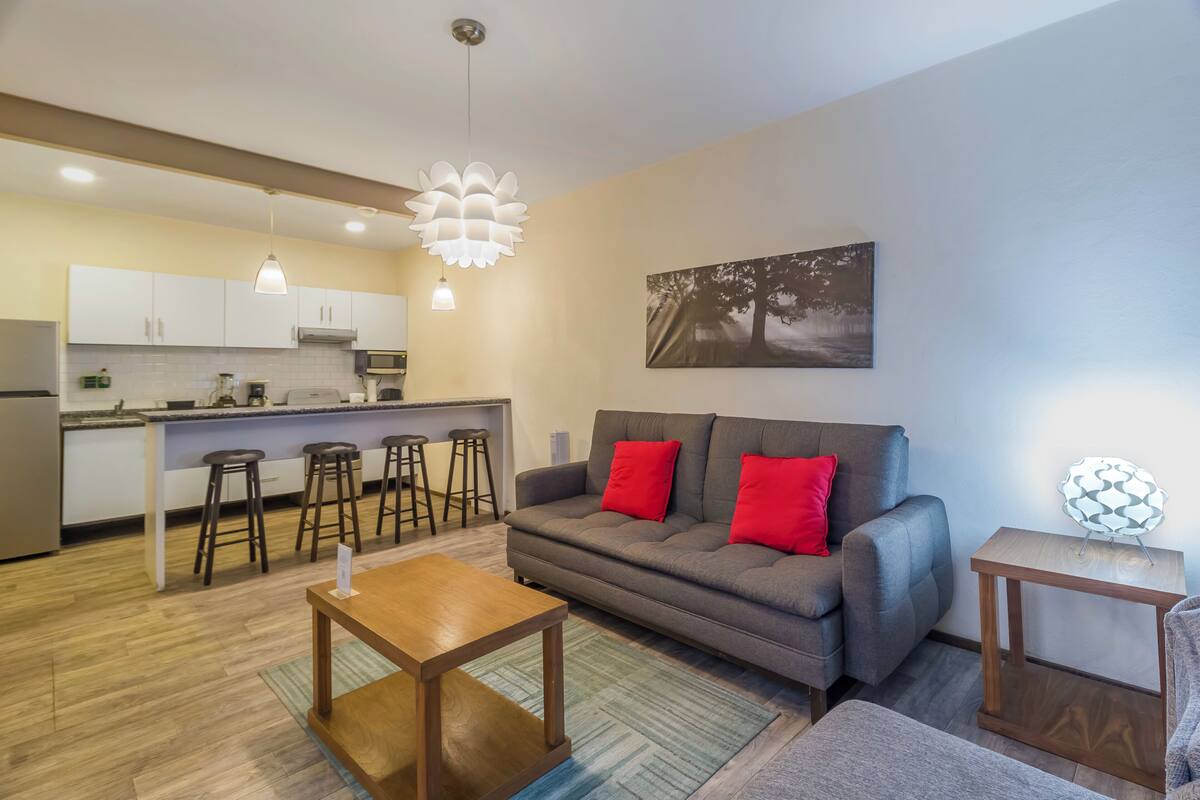 Un salon moderne avec un canapé gris, des coussins rouges, une table basse en bois et un coin cuisine doté de meubles blancs et de tabourets de bar.