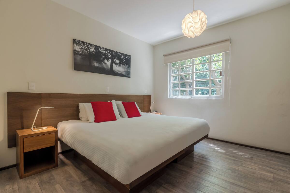 Une chambre moderne avec une tête de lit en bois, un grand lit avec une literie blanche et rouge, une table de chevet avec une lampe, une fenêtre avec des stores et un tableau accroché au mur.