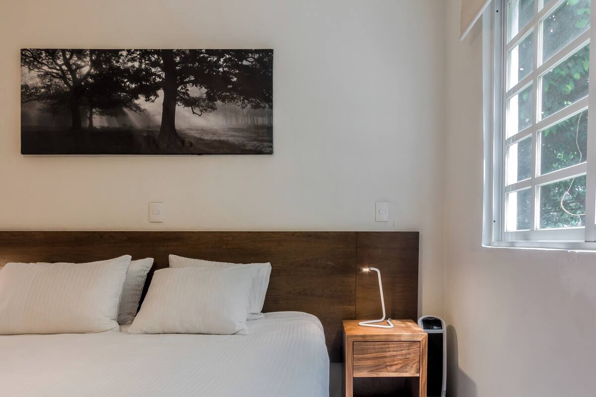 Une chambre à coucher avec un lit, une table de chevet, une lampe et une fenêtre donnant sur des arbres.