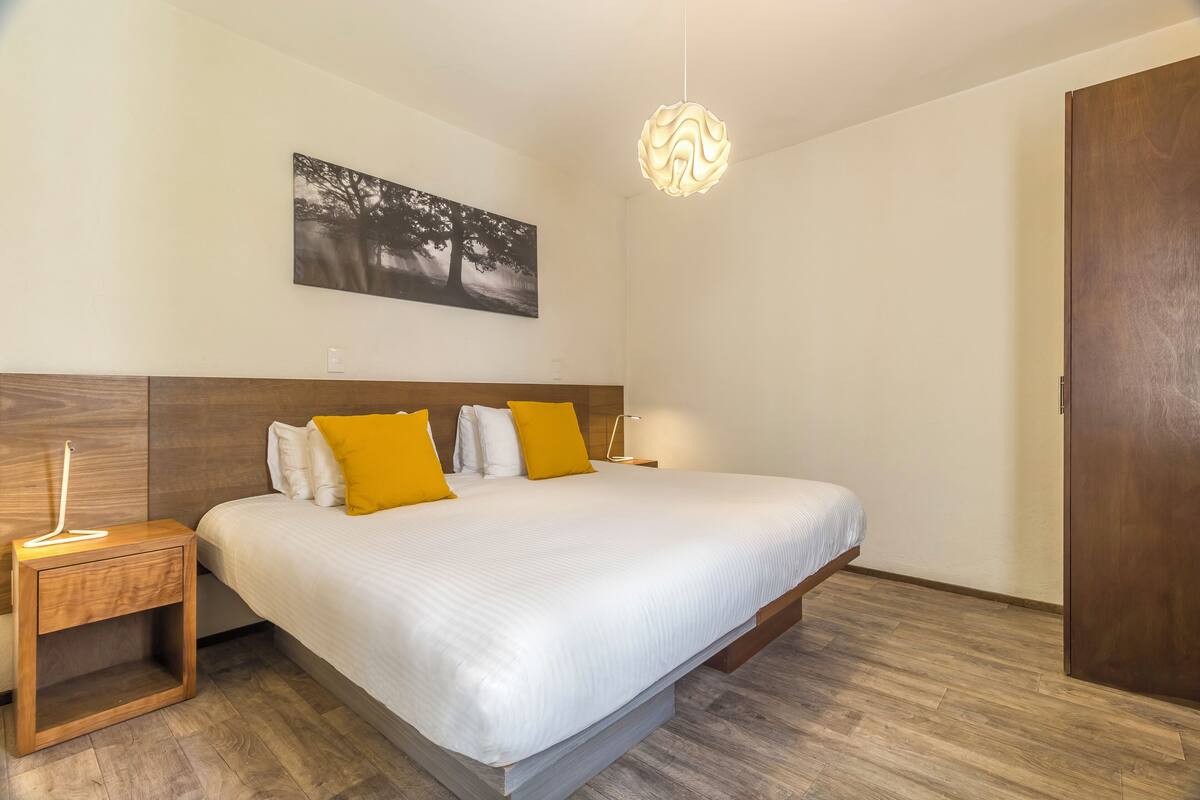 Une chambre à coucher avec un grand lit, une tête de lit en bois, une table de chevet avec une lampe, un tableau accroché au mur et une porte en bois.