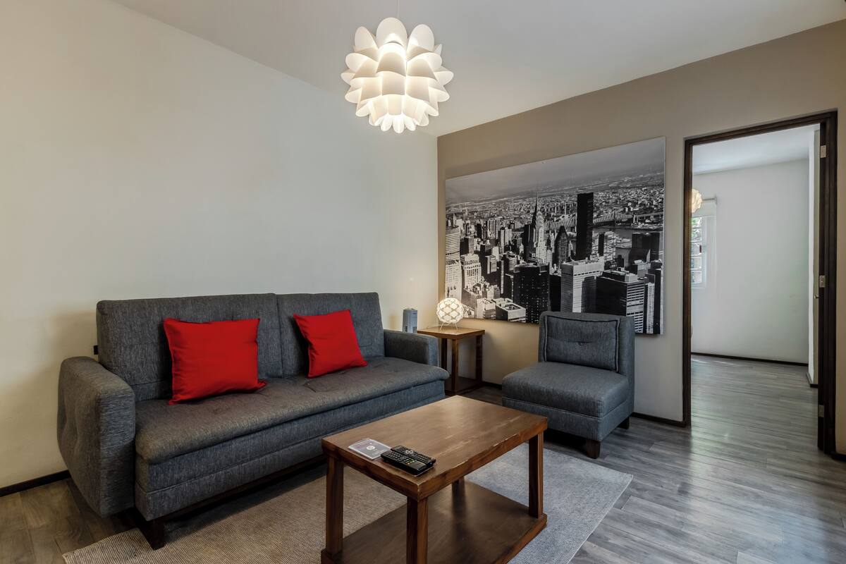 Un salon moderne avec un canapé gris, un coussin rouge, une table basse en bois et un tableau représentant un paysage urbain accroché au mur.