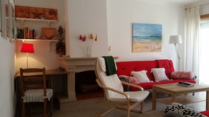 A living room with a red sofa, a wooden chair, a small table, a fireplace, and a painting of a beach scene. - Apartment sunny beach (Peniche)