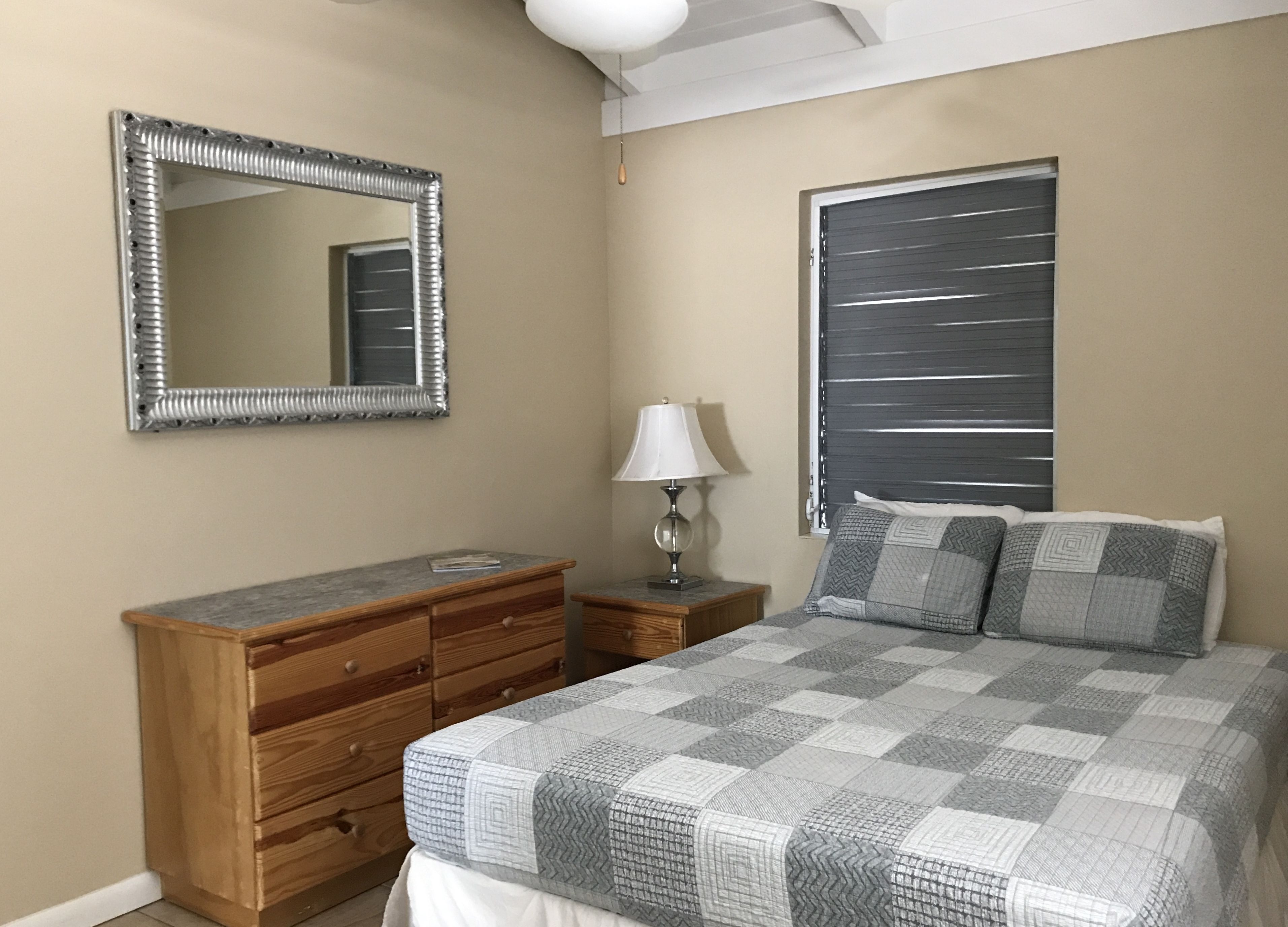 A bedroom with a bed, a wooden dresser, a mirror, a nightstand, a lamp, and a window with blinds.
