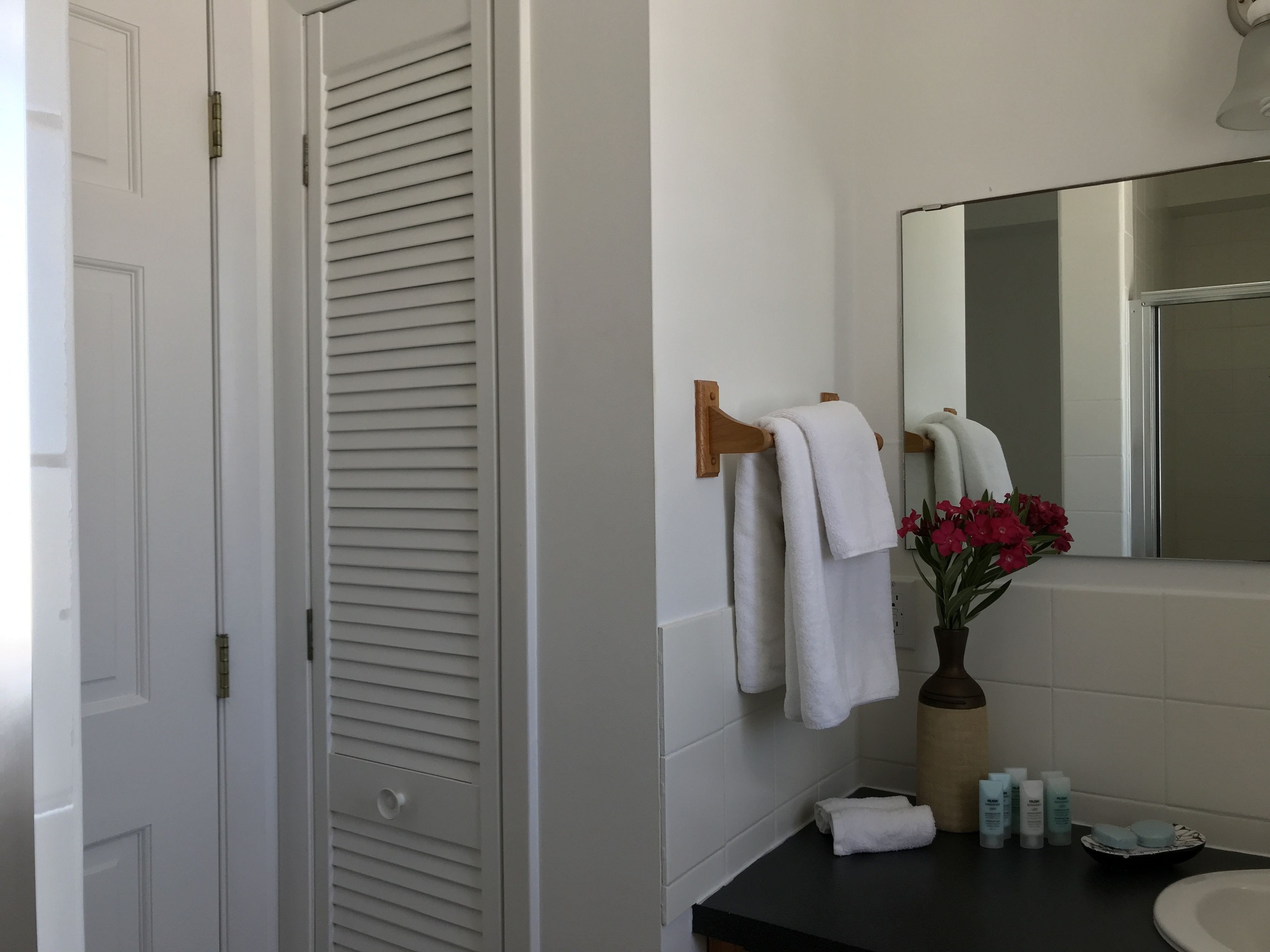 Une salle de bain avec une porte blanche, un porte-serviettes avec deux serviettes blanches, un miroir, un vase avec des fleurs rouges et des articles de toilette sur le comptoir.