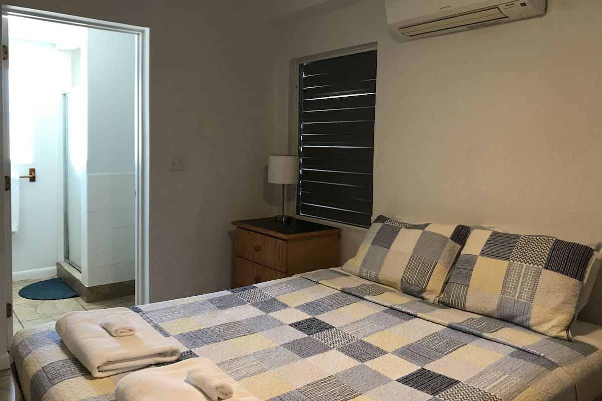 Un dormitorio con edredón a cuadros, lámpara de noche, ventana con persianas y equipo de aire acondicionado.