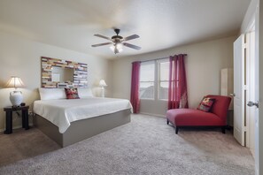Une chambre à coucher comprenant un lit, un ventilateur de plafond, une fenêtre avec des rideaux, une chaise et une table de chevet avec une lampe.