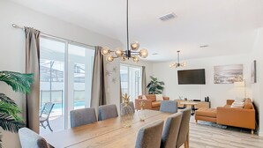 A modern living room with a wooden dining table, grey upholstered chairs, a brown sofa, and a TV mounted on the wall. - 🏝️ 7BR/5BA Home w/ Private Pool & Spa, Themed Kids’ Rooms & Resort Fun | Storey Lake Near Disney (Kissimmee)