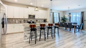 A modern kitchen with white cabinets, a granite countertop, and a built-in microwave. There are four bar stools with wooden seats and black metal legs. The kitchen has a large island with a sink and a stove. - 🏝️ 7BR/5BA Home w/ Private Pool & Spa, Themed Kids’ Rooms & Resort Fun | Storey Lake Near Disney (Kissimmee)