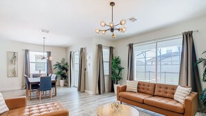 A spacious living room with a brown leather sofa, a round coffee table, and a dining area with a table and chairs. - 🏝️ 7BR/5BA Home w/ Private Pool & Spa, Themed Kids’ Rooms & Resort Fun | Storey Lake Near Disney (Kissimmee)