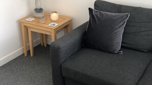 A dark grey sofa with a single black pillow, a small wooden side table with a white lamp, a lit candle, and two cards.
