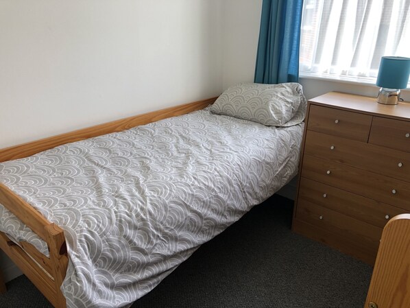 A single bed with a patterned bedspread, a wooden dresser with drawers, and a window with curtains.
