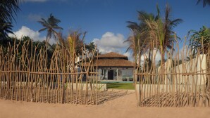 Une maison de plage avec un toit en tuiles, entourée de palmiers et délimitée par une clôture en bambou.