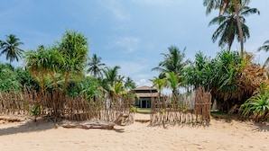 Une plage bordée d’une clôture en chaume, avec des palmiers et un bâtiment doté d’un toit en tuiles.
