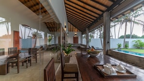 Un espace intérieur vaste et lumineux, avec un plafond en bois, une table à manger entourée de chaises, un coin salon agrémenté de coussins et une vue sur une piscine à l’extérieur.