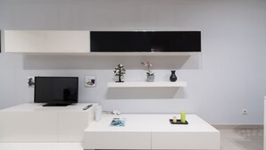 A modern living room with a flat-screen TV, white furniture, a small table with a magazine, and decorative items on shelves.