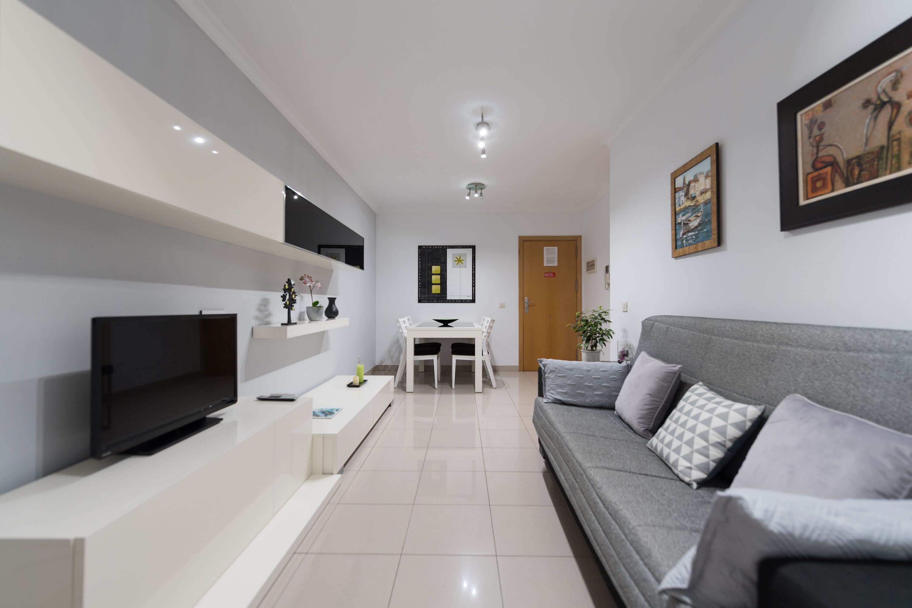 A modern living room with a flat-screen TV, a grey sofa, and a dining area with a white table and chairs.