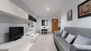 A modern living room with a flat-screen TV, a grey sofa, and a dining area with a white table and chairs.