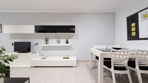 A modern living room with a flat-screen TV, white furniture, and a dining area with white chairs.