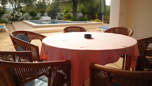 A round table with a pink tablecloth surrounded by wicker chairs, a small black pot, and a pool in the background.
