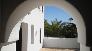 An arched corridor with white walls and a tiled floor, leading to a palm tree and a clear blue sky.