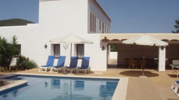A pool area with lounge chairs and umbrellas, a white building, and a clear sky.