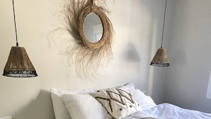 Une chambre à coucher comprenant un lit, deux tables de chevet, une petite table, une décoration murale en fibres tressées et deux lampes suspendues.