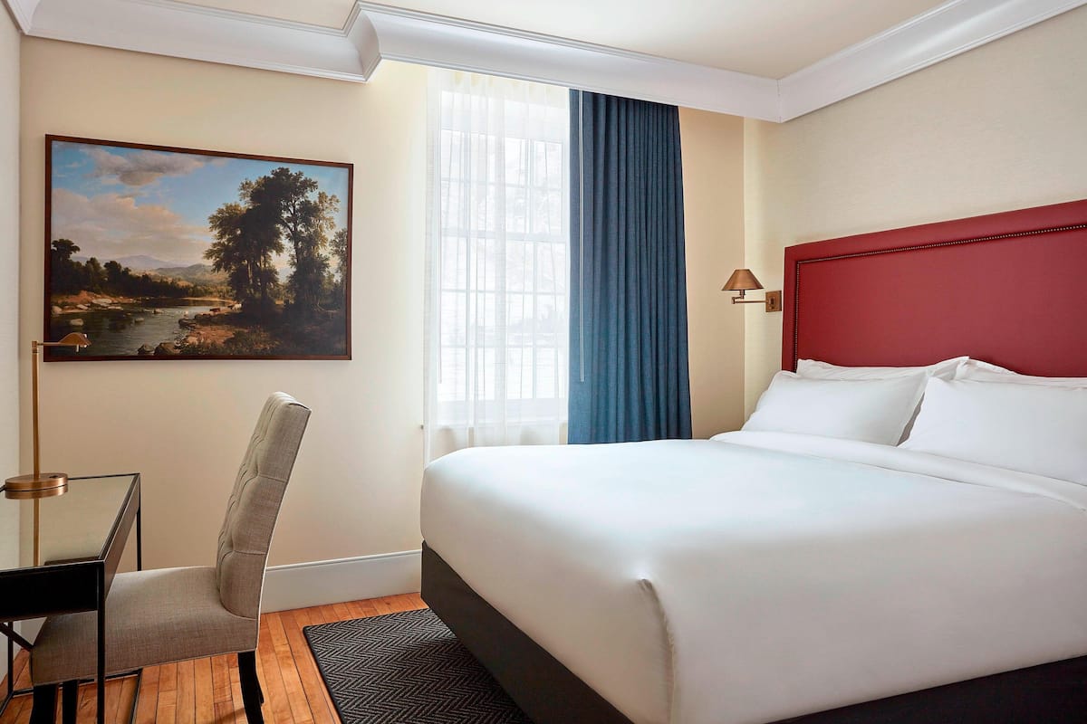 Habitación de hotel con una cama grande, un escritorio, una silla, un cuadro de un paisaje y una ventana con cortinas.