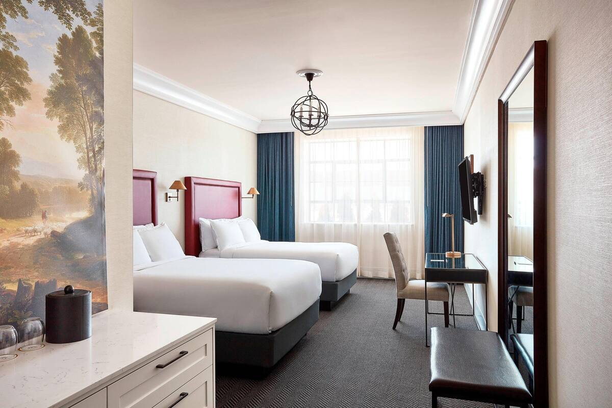 Habitación de hotel con dos camas, un escritorio, una silla, un espejo grande y un cuadro en la pared.