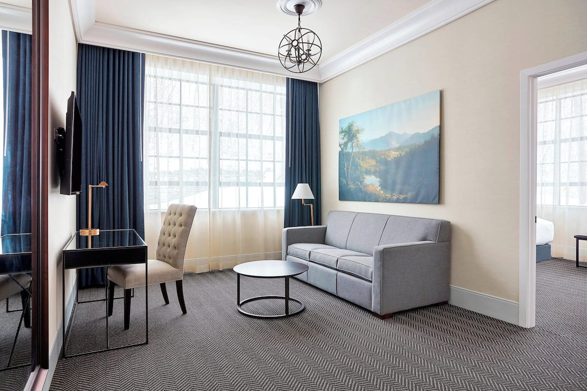 Habitación de hotel moderna con sofá gris, una mesita redonda, televisor montado en la pared y un ventanal con cortinas azules.