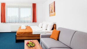 A hotel room with a bed, a gray sofa, a wooden coffee table with fruit, and a painting on the wall. - Apartmánový dom Família Smokovec (Vysoké Tatry)