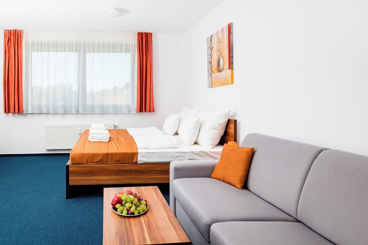 Une chambre d’hôtel avec un lit, un canapé gris, une table basse en bois sur laquelle se trouvent des fruits, et un tableau accroché au mur.
