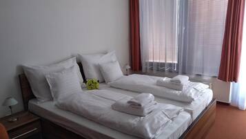 Una cama doble con ropa de cama blanca, una mesita de noche con lámpara y una ventana con cortinas.