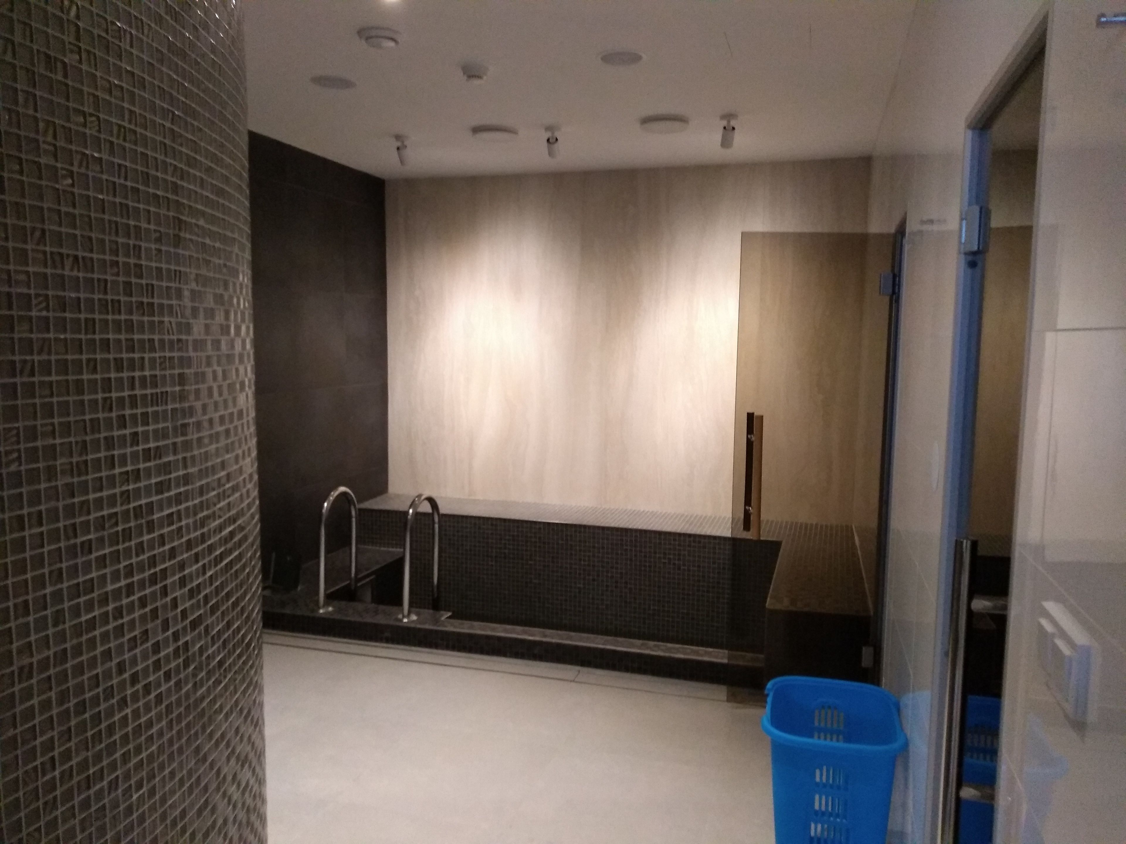 Een moderne badkamer met een glazen douchecabine, een donkere betegelde muur en een blauwe plastic mand op de vloer.