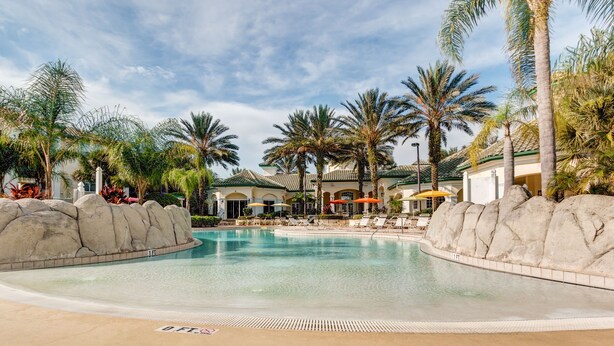 A pool area with lounge chairs and umbrellas, surrounded by palm trees and a building with a tiled roof. - Beautiful Lakeside Penthouse Award Winning Resort Just Minutes to Disney! (Kissimmee)