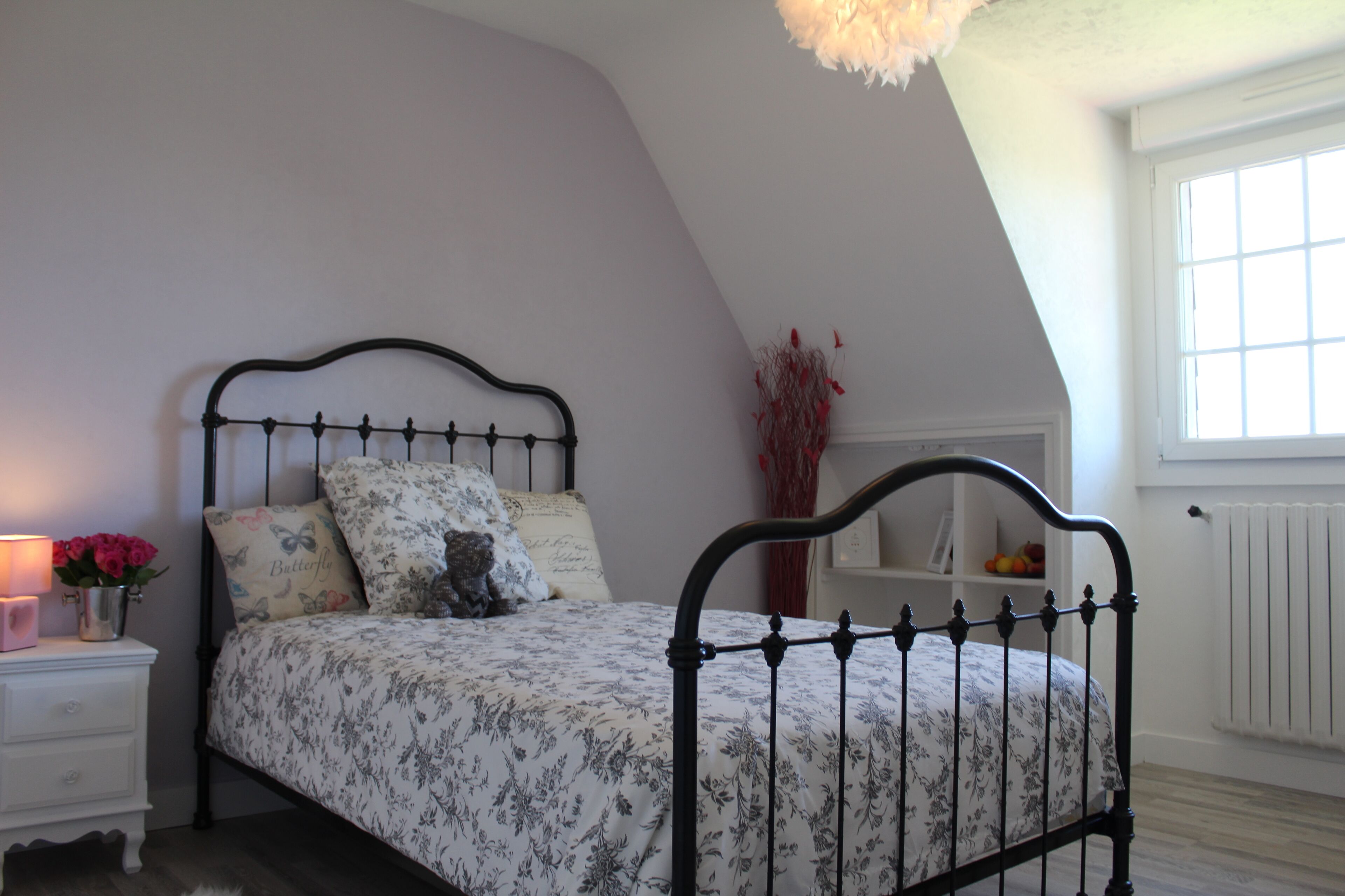 A bedroom with a bed, a nightstand, a vase with flowers, a radiator, and a window.