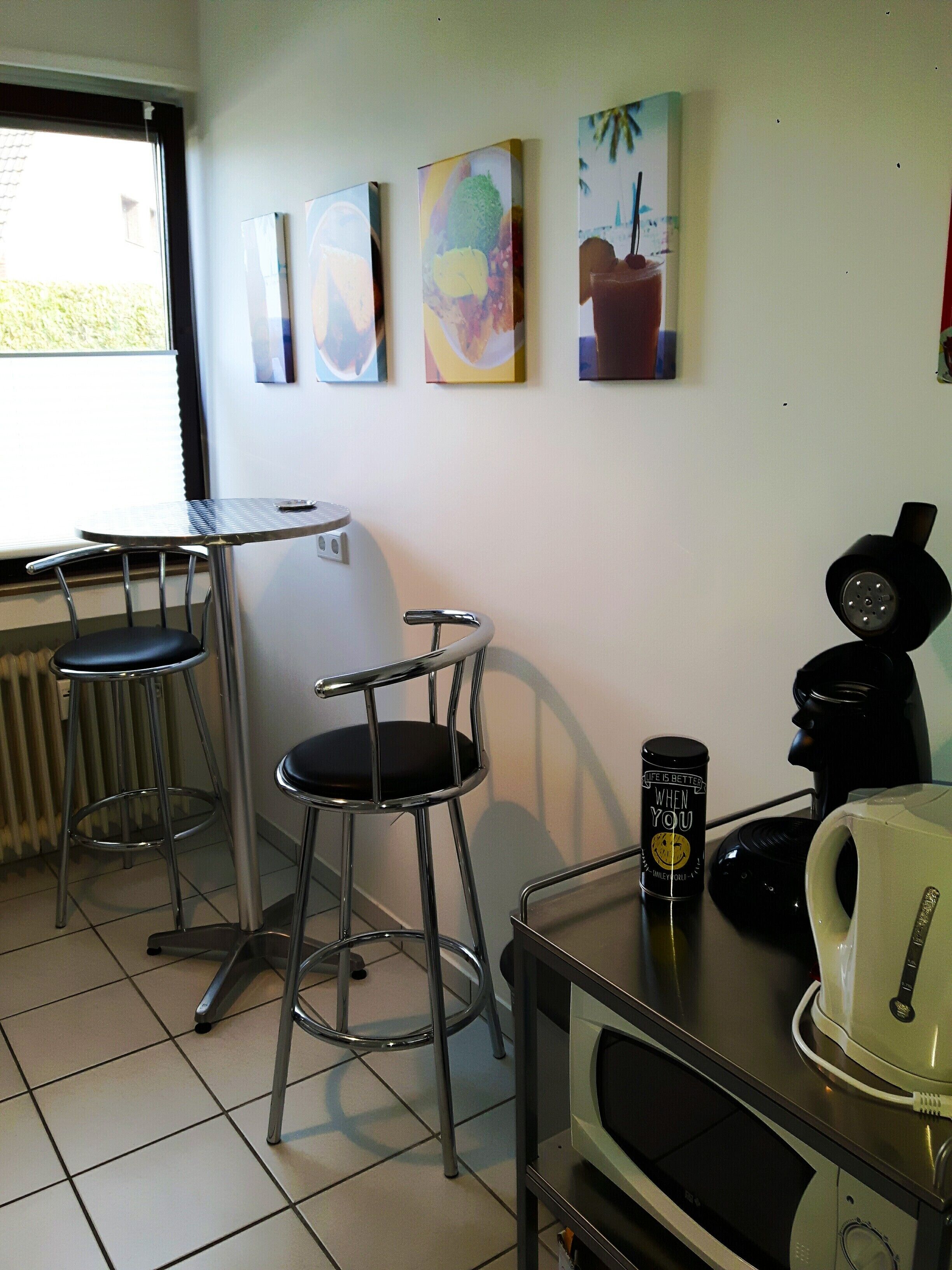 Uma pequena cozinha equipada com cafeteira, chaleira e micro-ondas. Há duas banquetas e uma janela com persianas.