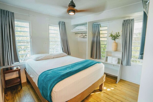 Un dormitorio con cama, mesita de noche, ventilador de techo y ventana con cortinas.