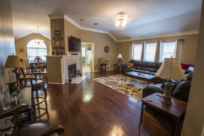 A spacious living room with hardwood flooring, a fireplace, a dining area with chairs and a table, and a comfortable seating area with a sofa and lamps. - Great Location! Only 5-10 minutes from Kyle Field! (Bryan)