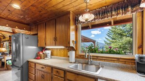 A kitchen with wooden cabinets, a stainless steel refrigerator, a coffee maker, and a window with a view of greenery and mountains.