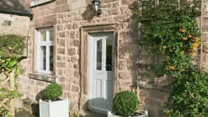 A stone building with a white door, two windows, and a small porch area with potted plants.