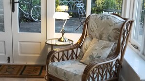 A corner room with a wicker chair, a small round table with a lamp, and a view of bicycles outside.