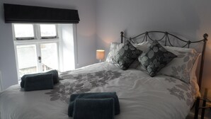 A neatly made bed with patterned pillows and folded towels, a bedside lamp, and a window with blinds.