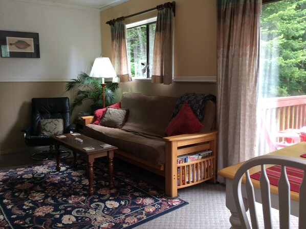 Ein gemütliches Wohnzimmer mit einem braunen Sofa, einem Holztisch, einem schwarzen Sessel und einem roten Stuhl. Ein Fenster mit Vorhängen, eine Lampe und ein Bücherregal mit Büchern sind ebenfalls vorhanden.
