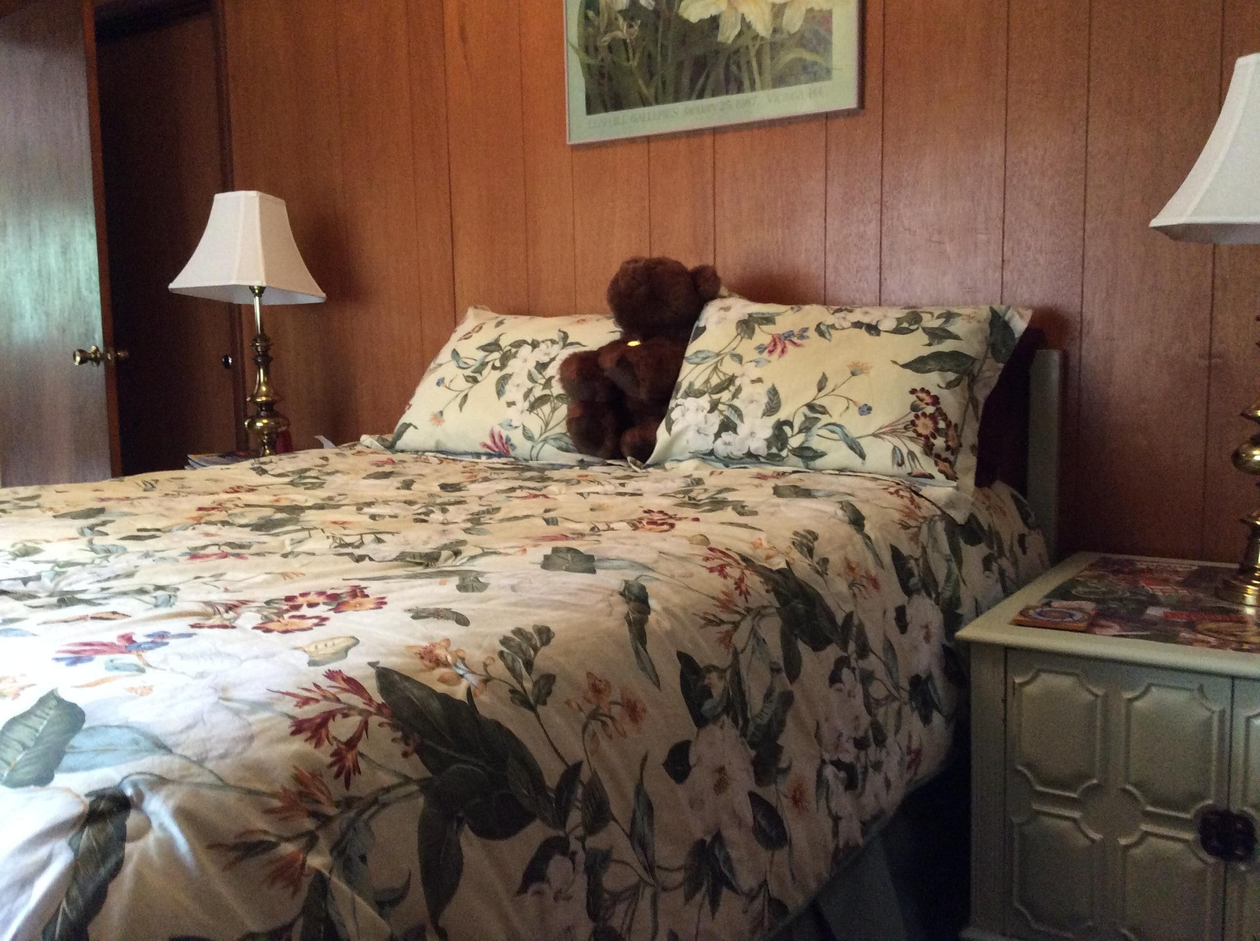 A bed with floral bedding, a teddy bear on the pillow, and a nightstand with a lamp.