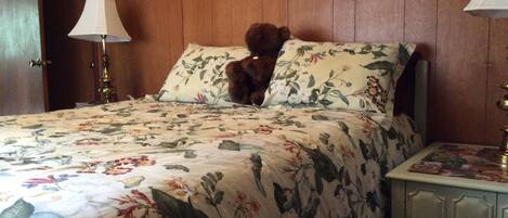 A bed with floral bedding, a teddy bear on the pillow, and a nightstand with a lamp.