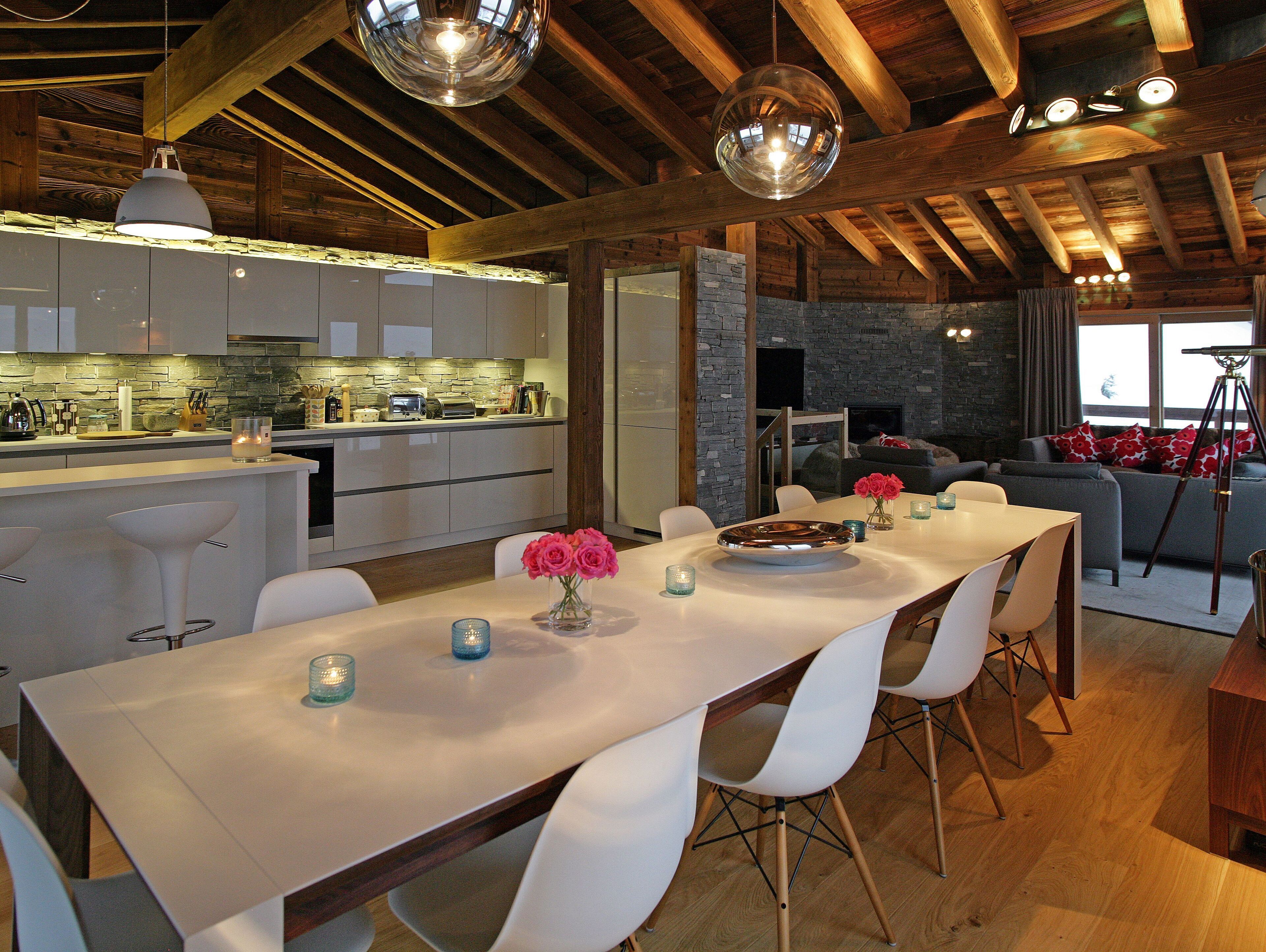 Een moderne keuken met een achterwand van steen, houten balken en een eetgedeelte met witte stoelen en een centrale tafel.