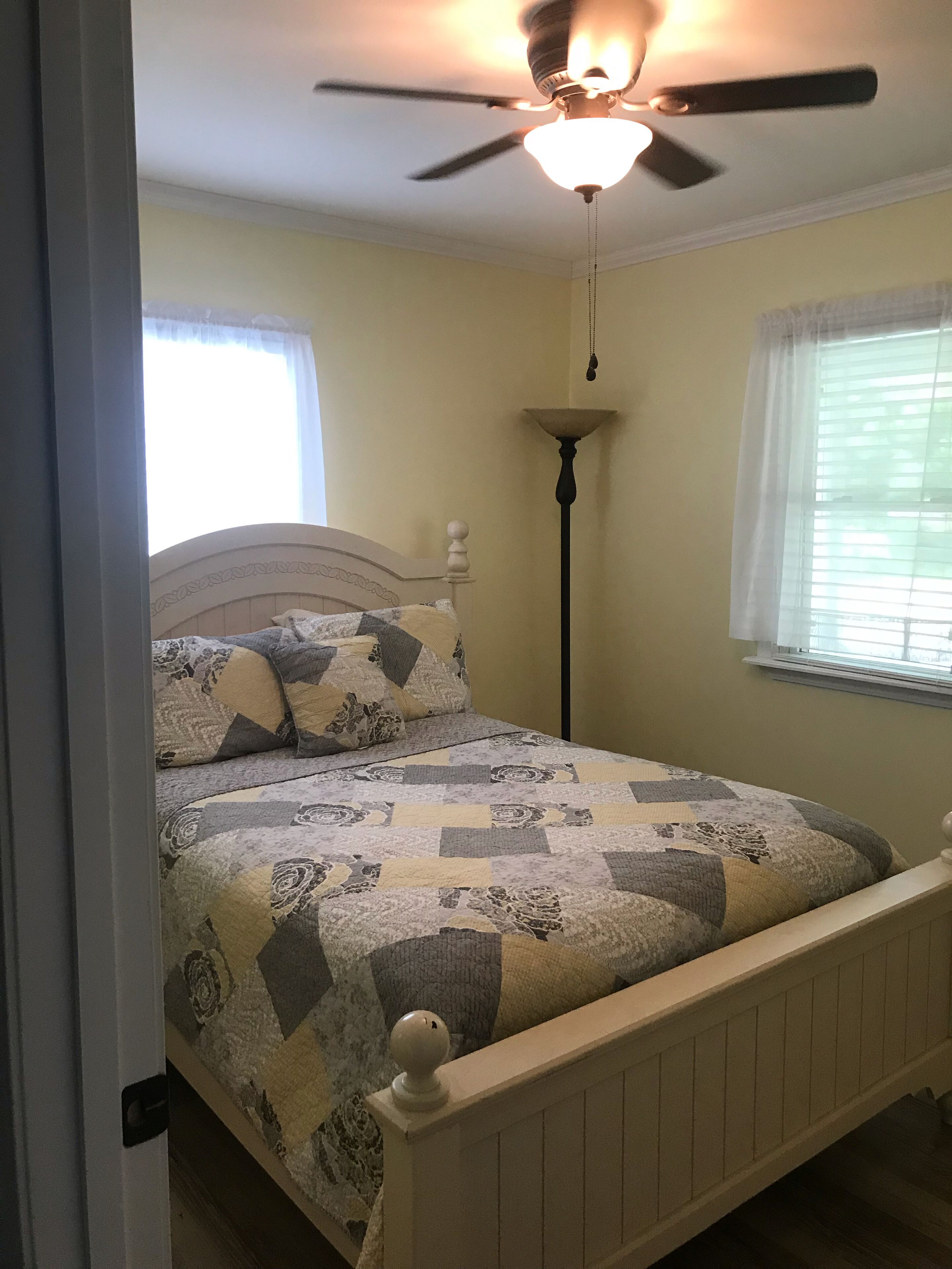 A bedroom with a bed, a ceiling fan, a floor lamp, and windows with blinds.